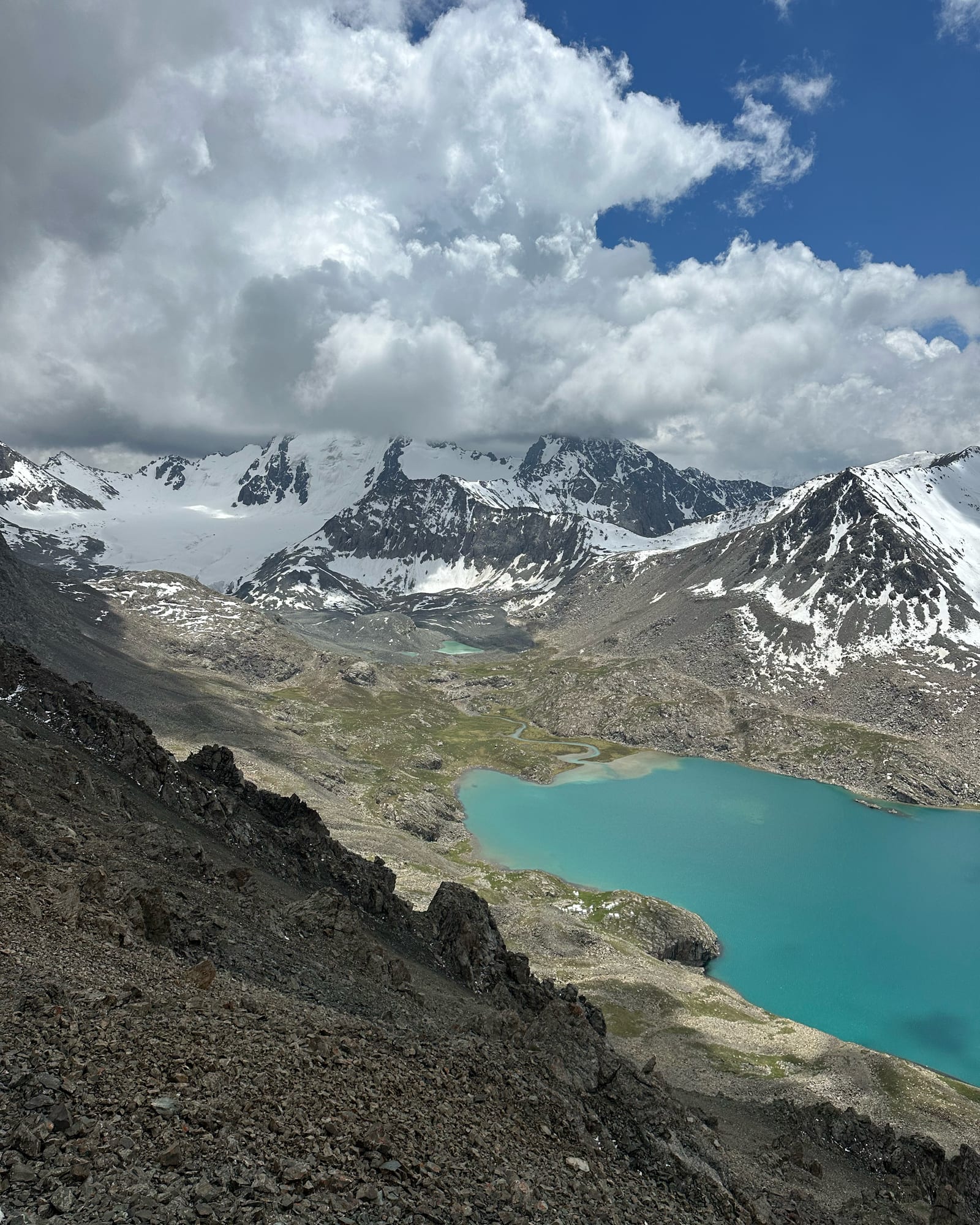 Ledovcové jezero Ala-Kul pod zasněženými štíty a mraky, v popředí suťový svah