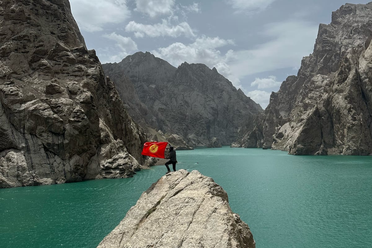 Postava s kyrgyzskou vlajkou na skalním ostrohu nad tyrkysovým jezerem Kel-Suu